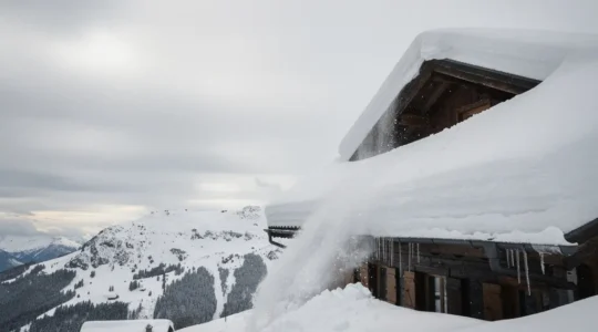Chalet de montagne avec une toiture très pentue couverte de neige, une coulée de neige en cours et un grand ciel dégagé laissant de l’espace libre.